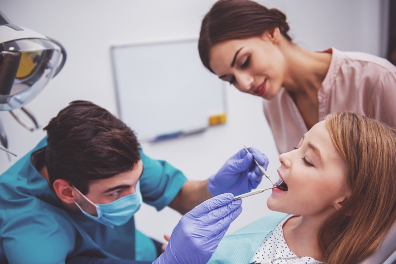 Pediatric dentist examining young patient with her mother in dental clinic