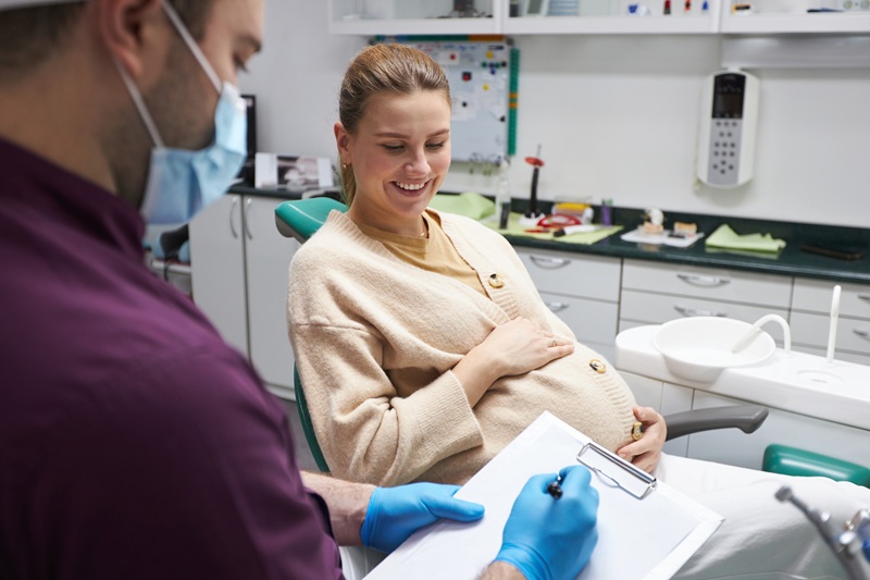 Dentist hygienist prescribing a dental prophylaxis treatment to a young pregnant woman, smiling while sitting on dentist's chair. The concept of a preventive examination by a dentist during pregnancy