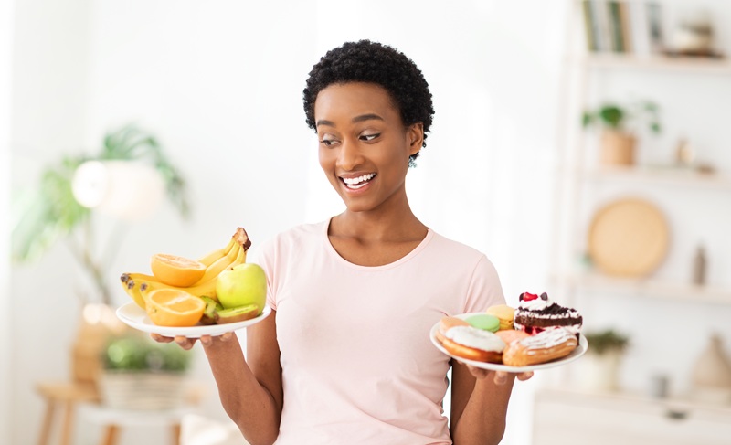 Best nutrition for slimming. Cheerful black woman holding plates with fruits and desserts indoors, choosing between healthy and junk foods, trying to keep balanced diet